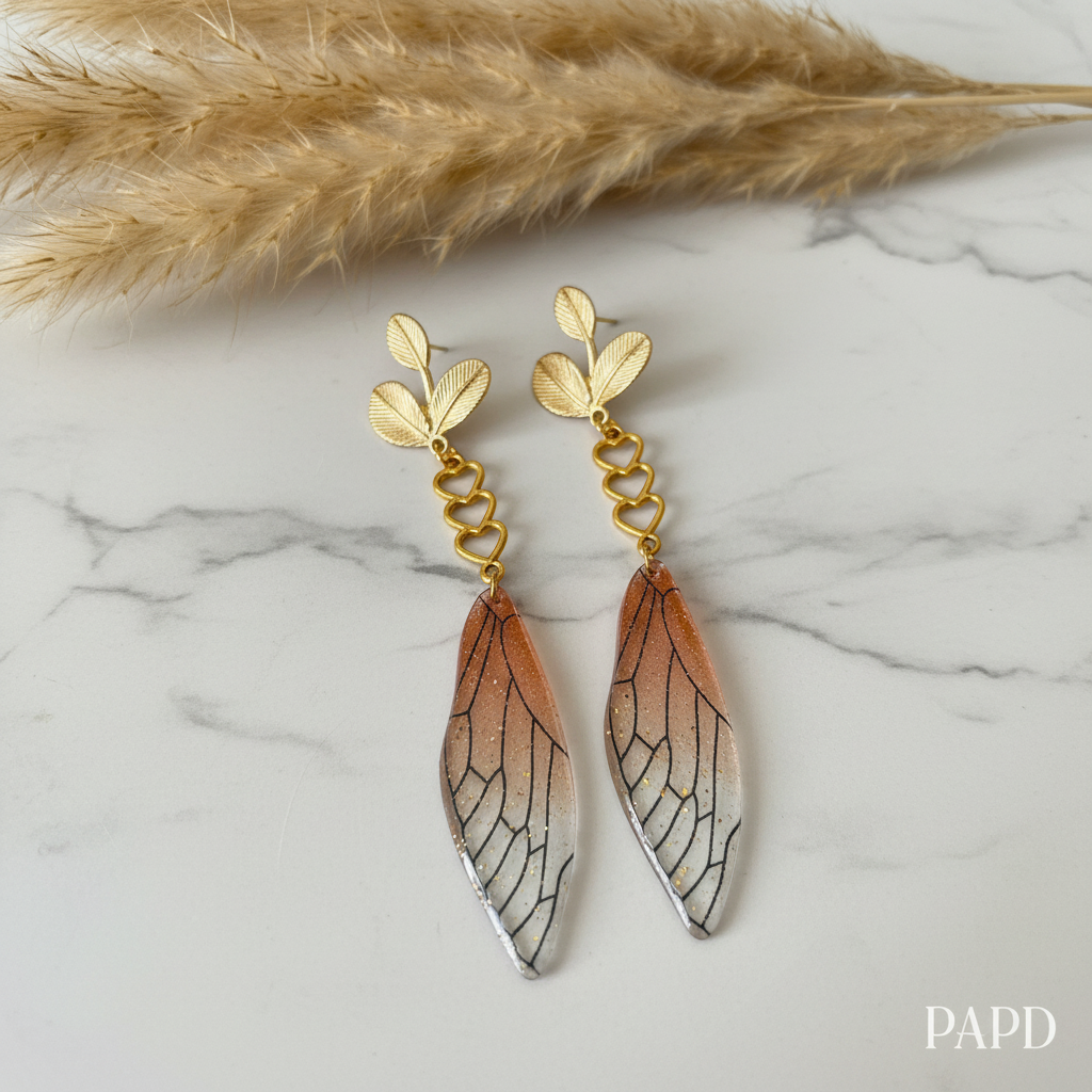 Boucles pendantes dorées avec ailes stylisées, cœurs et détails veinés brun-ocre.