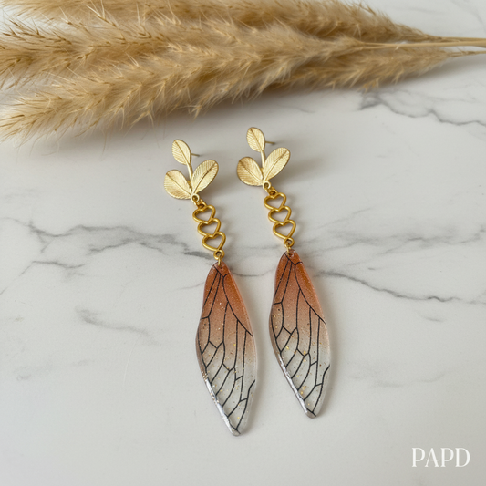 Boucles pendantes dorées avec ailes stylisées, cœurs et détails veinés brun-ocre.